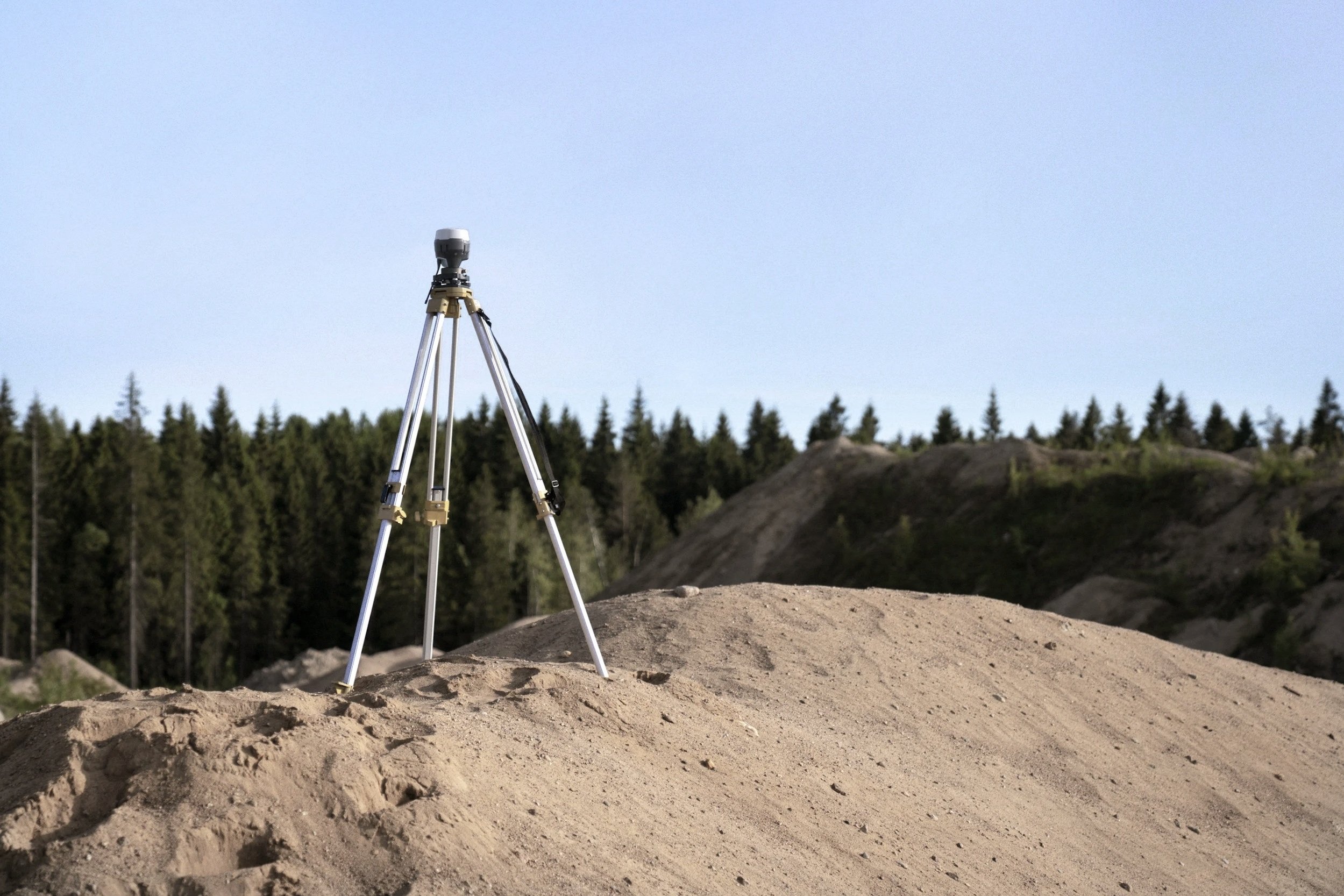 Surveying instrument on a tripod atop a gravel mound with forest behind